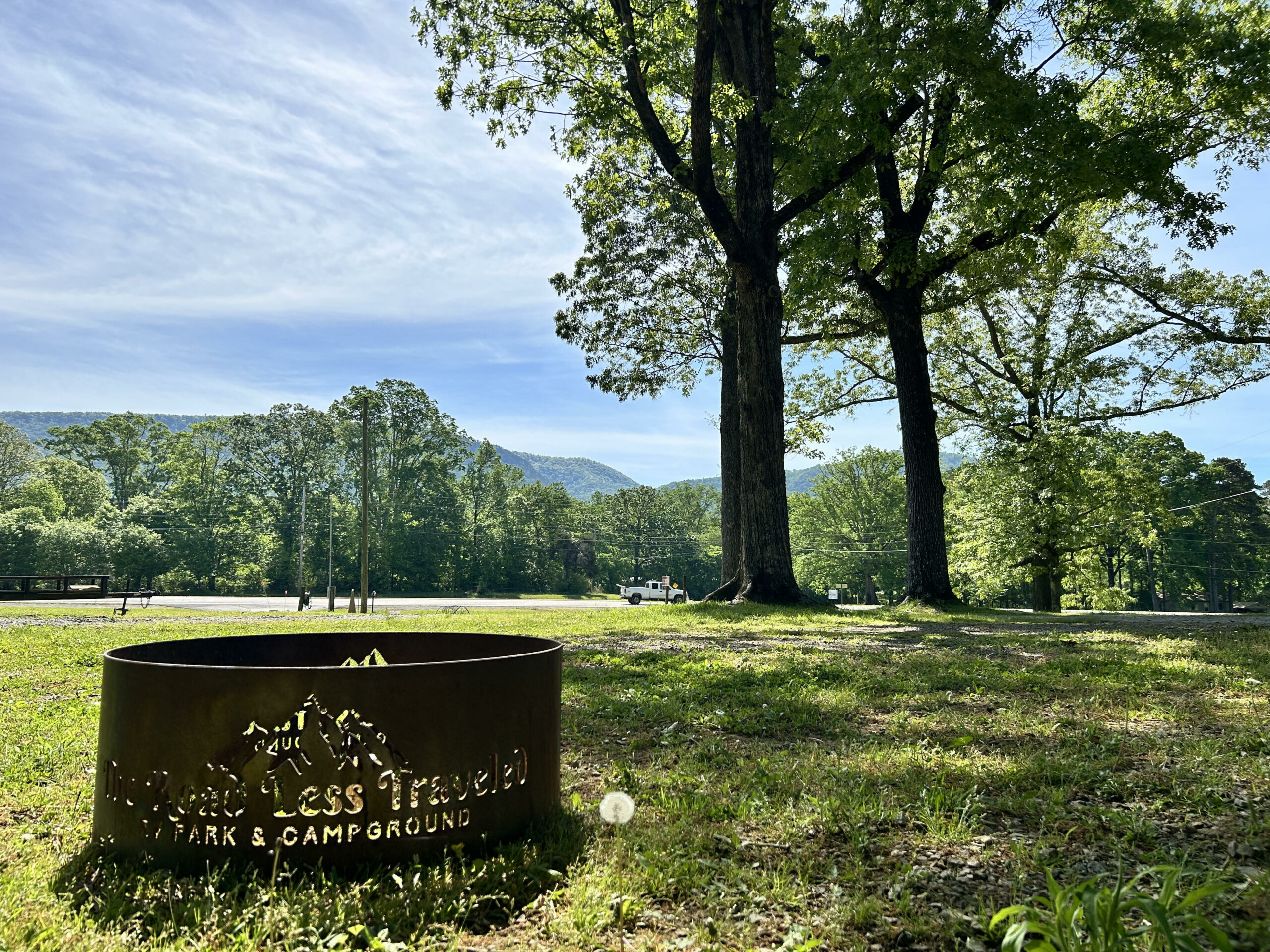 A metal fire pit sits in a grassy area under tall trees, with a view of distant mountains and a clear sky.
