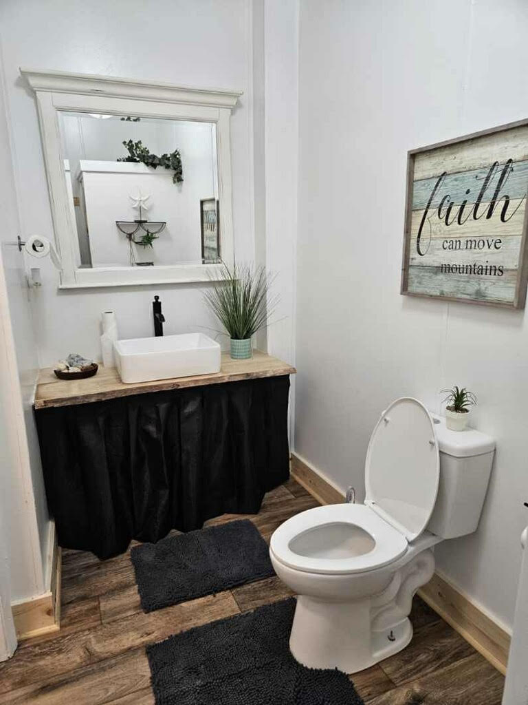A bathroom with a toilet, a sink on a wooden vanity, a mirror, potted plants, and a wall sign reading "faith can move mountains.